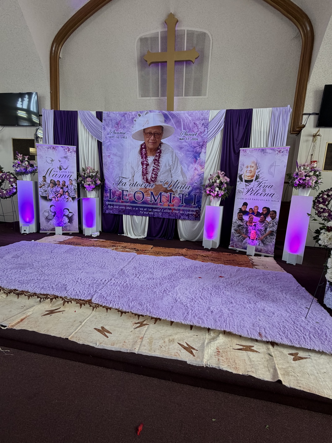 Purple and white memorial tribute with draped backdrop, portrait banners, floral arrangements and purple uplighting